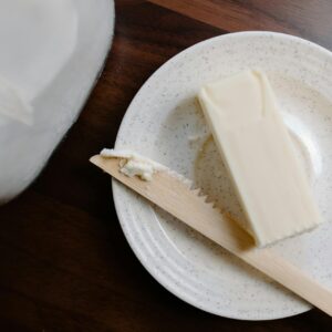 A stick of butter on a rustic plate with a wooden knife against a dark wooden surface.